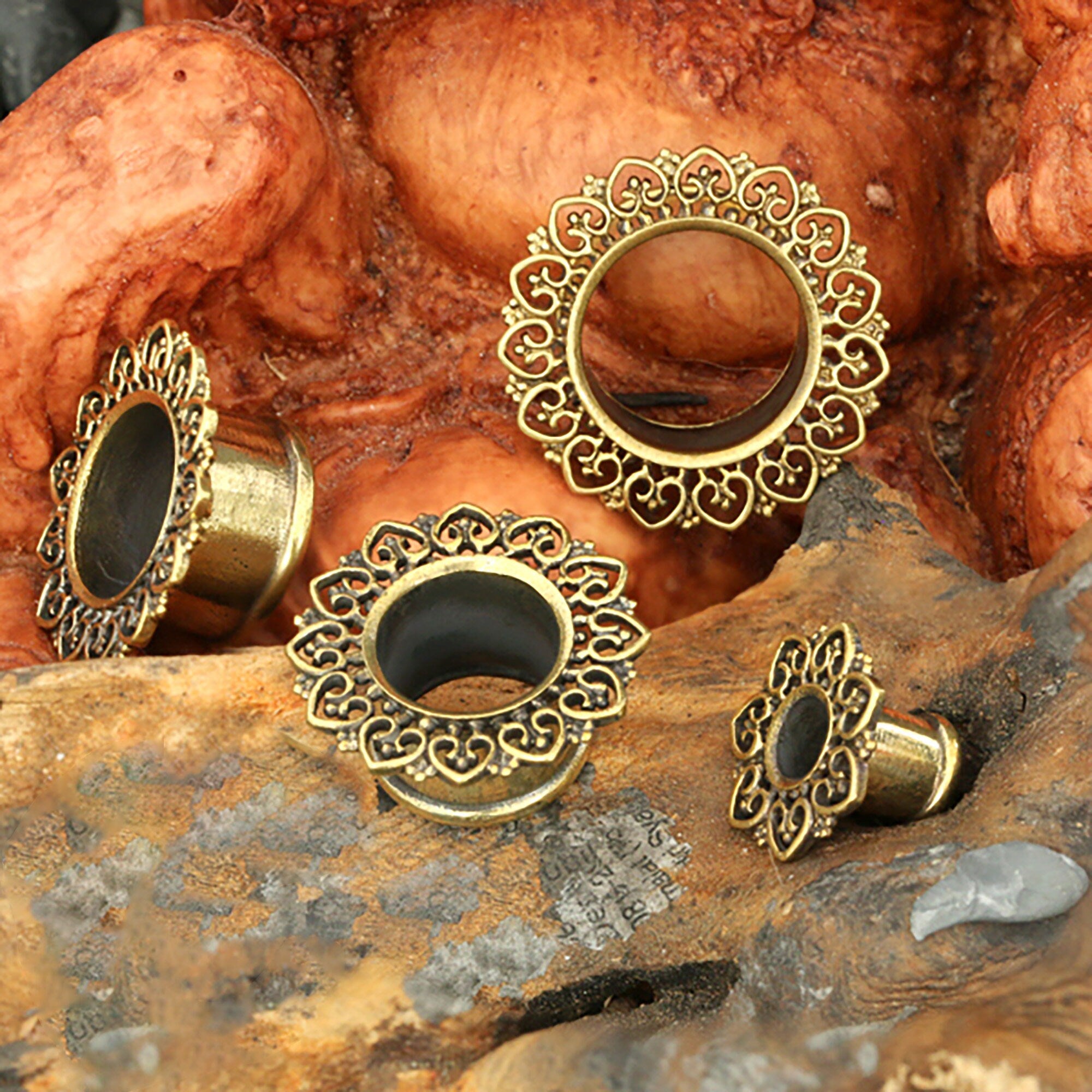A set of Indian Gold Brass Ear Gauges Tunnels Jewelry is displayed against a rustic, textured backdrop. These Ethnic Ear Plugs & Gauge Earrings from Kaya Movement feature intricate mandala designs in gold and come in varying sizes, showcasing elaborate craftsmanship with a vintage aesthetic. Perfect for body modification enthusiasts, these handmade Mandala Brass Tunnels are truly exquisite.